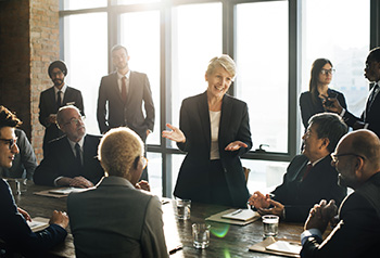 A team looking towards a mature woman standing and speaking during a meeting.