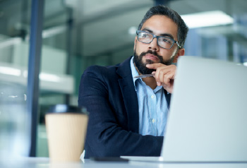 A person at work looking contemplative with a laptop and a coffee cup in the foreground.