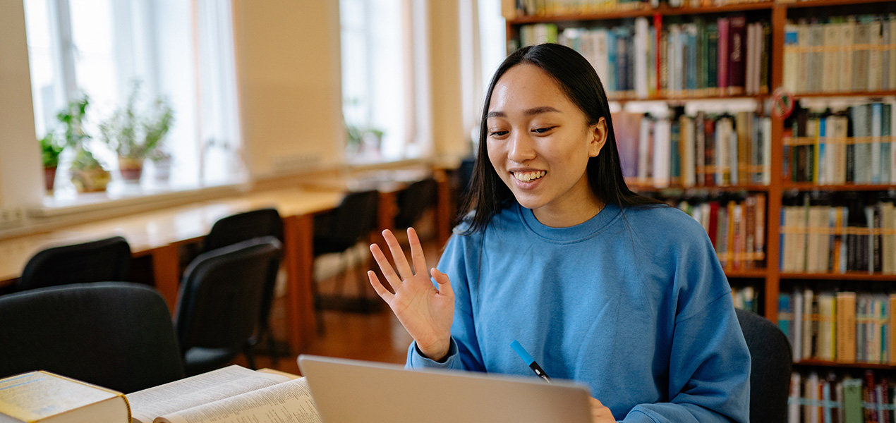 A young student surrounded by books starting a zoom meeting on a laptop.