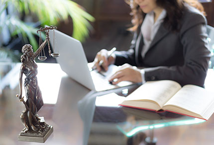 A statue of Lady Justice sits on a desk in the foreground, while in the background is a business woman working on a laptop.