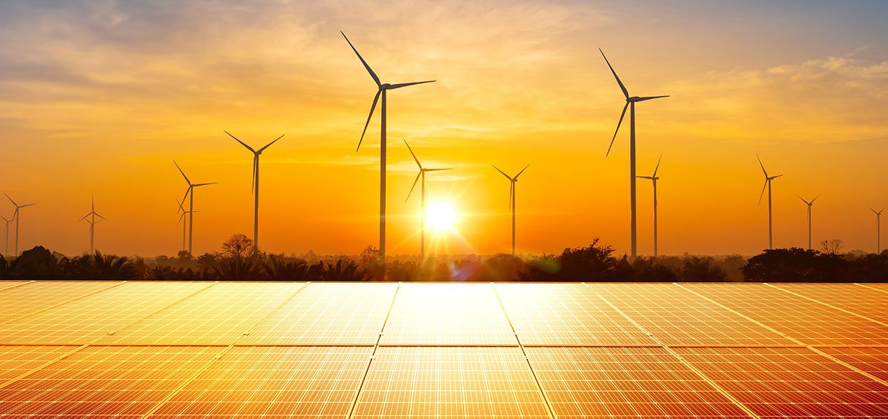 The sun setting behind a large set of solar panels and wind turbines in the distance.