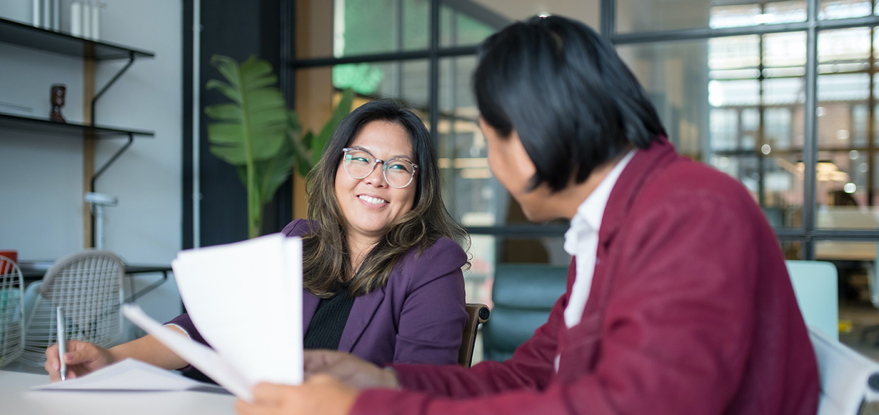 A smiling woman facing the camera sits with a man holding papers.