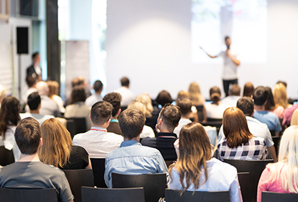 Back of an audience looking towards a speaker giving a talk at an event.