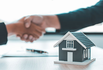 Two people shaking hands across a table that has papers and a model of a home on the table.