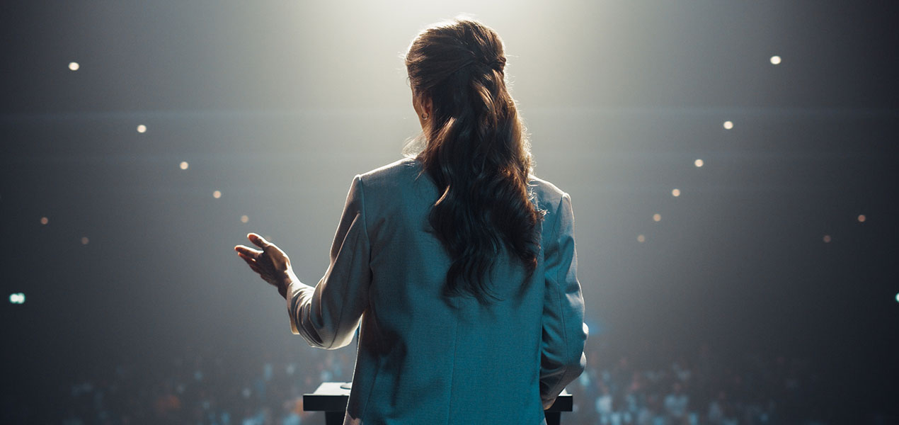 Back of an audience looking towards a speaker giving a talk at an event.