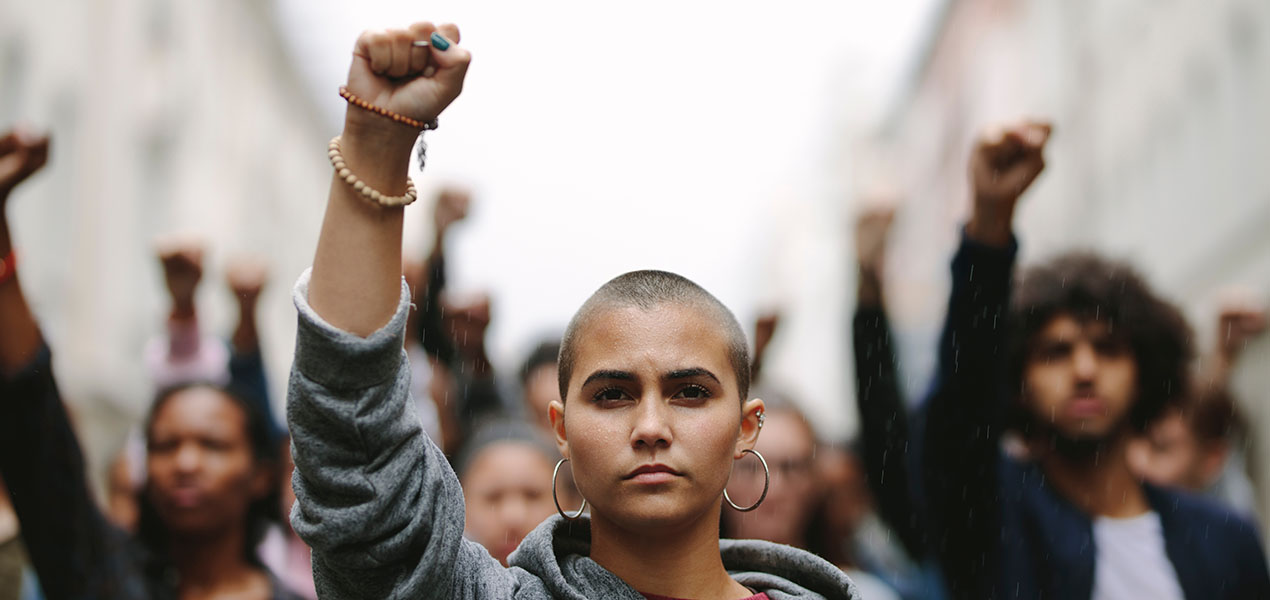A young protester with a raised fist stands in front, backed by a group also raising their hands.