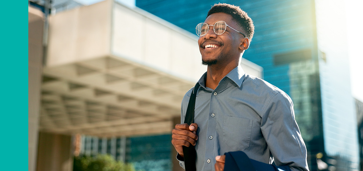 A young professional man walking in down a city street in the sun.