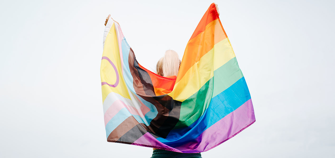 Back view of a person holding a rainbow Progress Pride flag behind their shoulders.