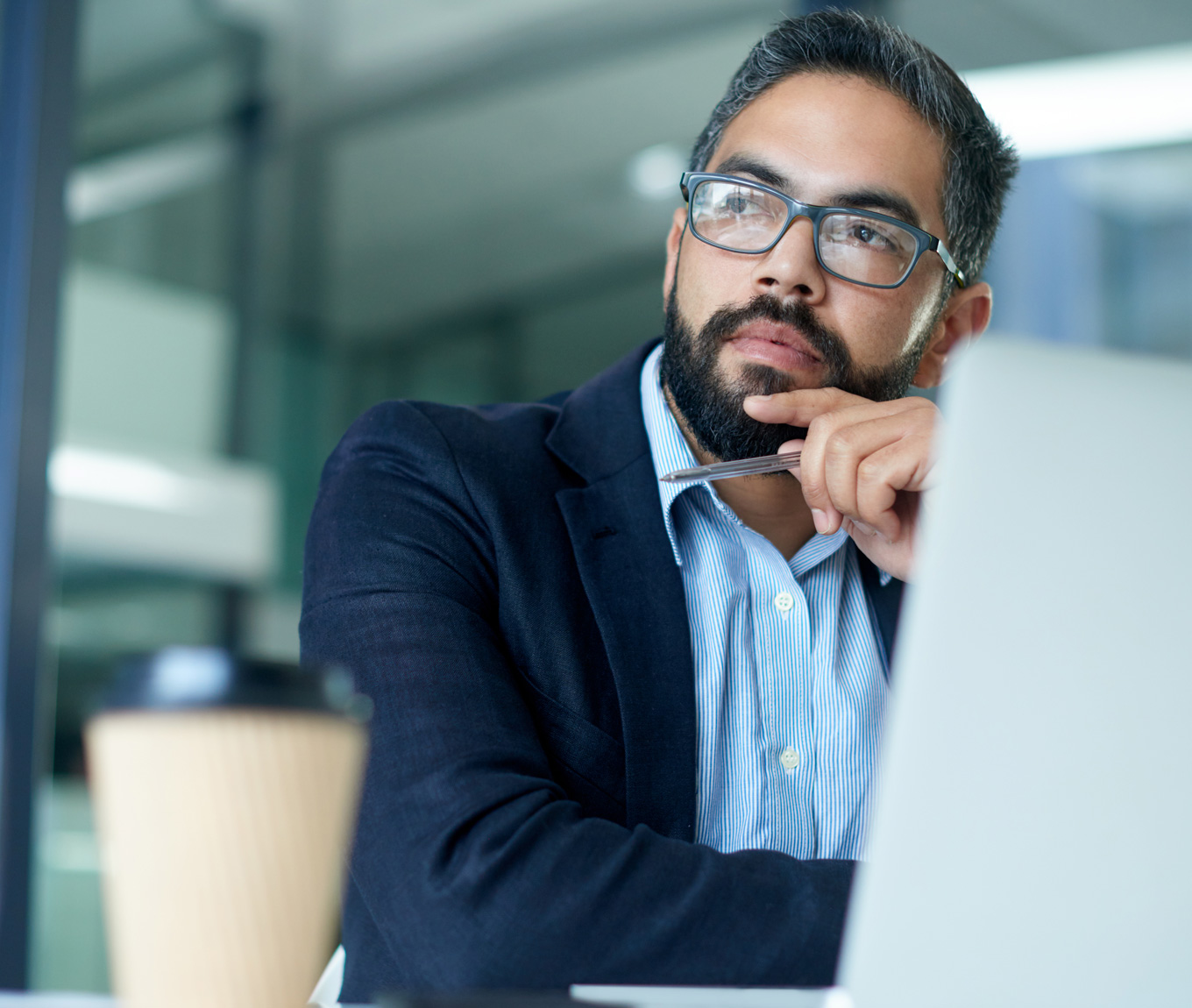 A person at work looking contemplative with a laptop and a coffee cup in the foreground.