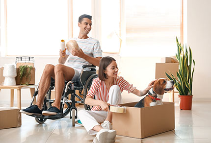 Young woman with her husband in wheelchair and dog packing things in a room on moving day.