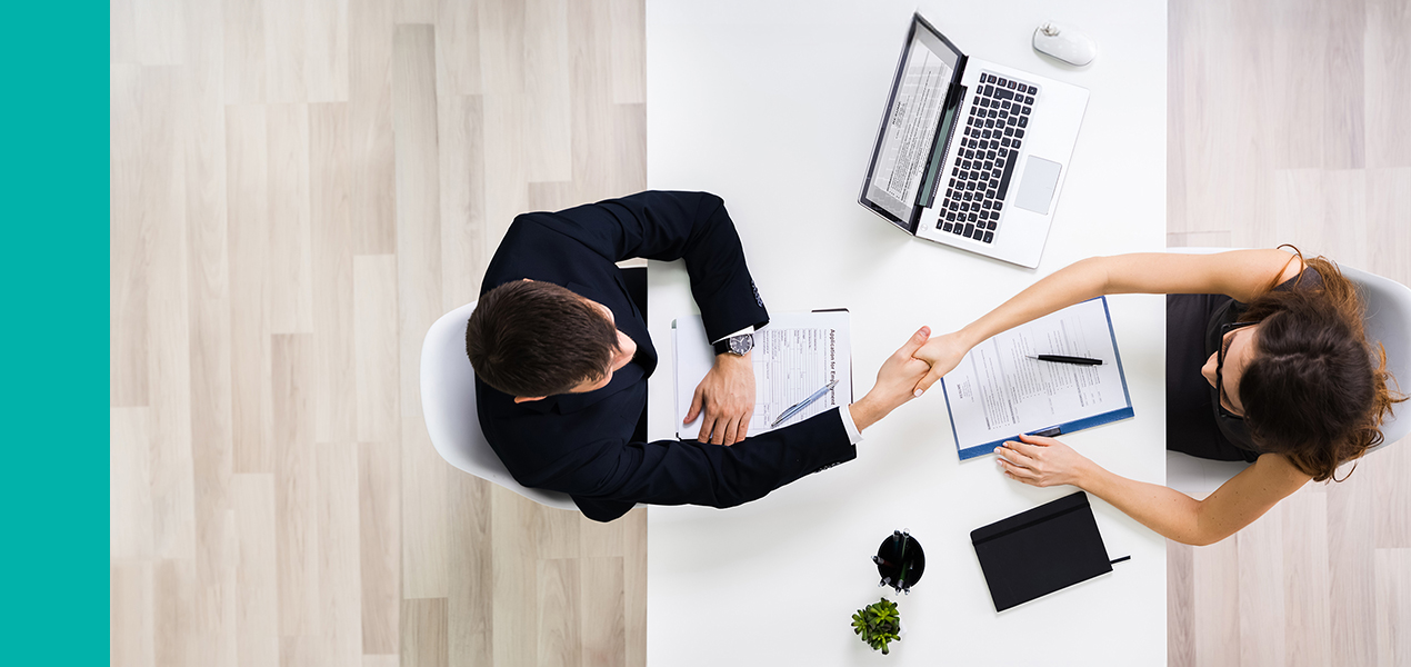 An aerial view of two professionals sitting at a desk shaking hands at the end of an interview.