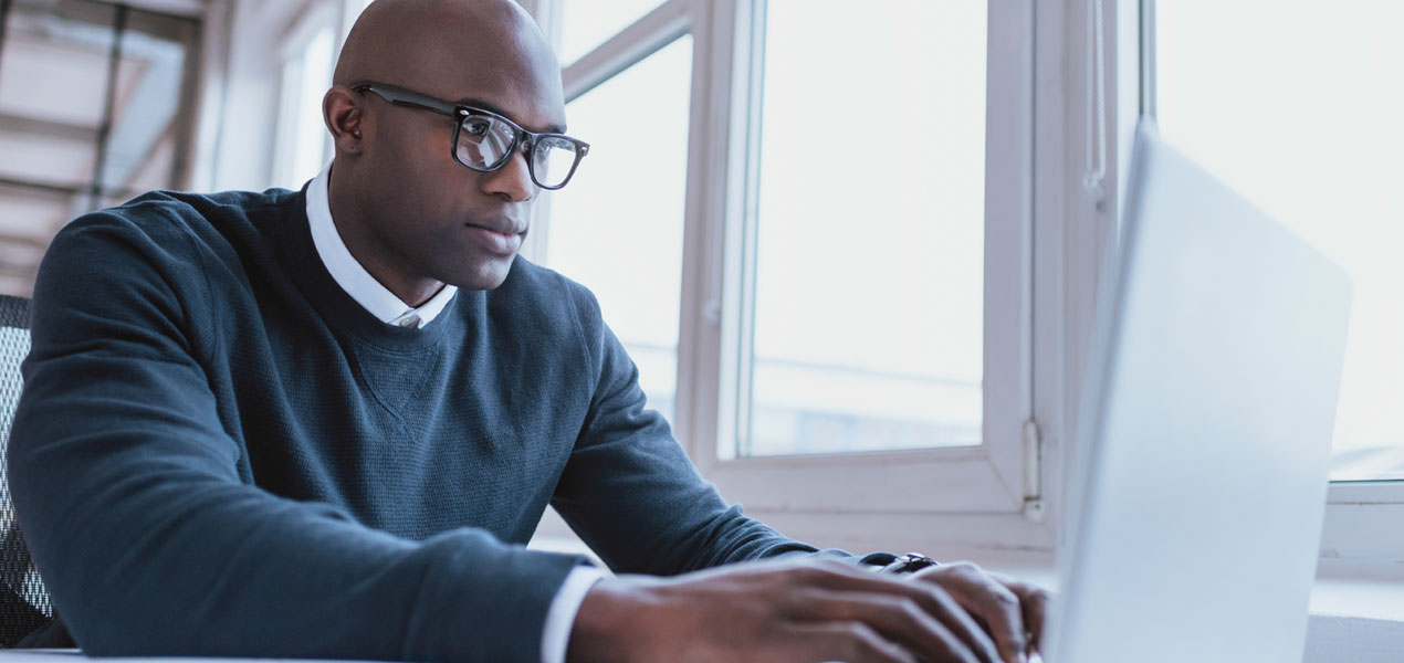 A Black business man working on a laptop by a large window.