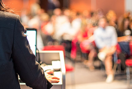 A presenter speaking at a podium in front of a large audience.