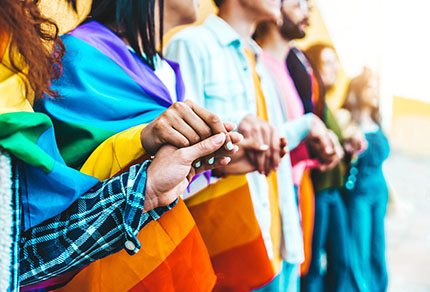 Group of people holding hands with rainbow flag draped across some participants.