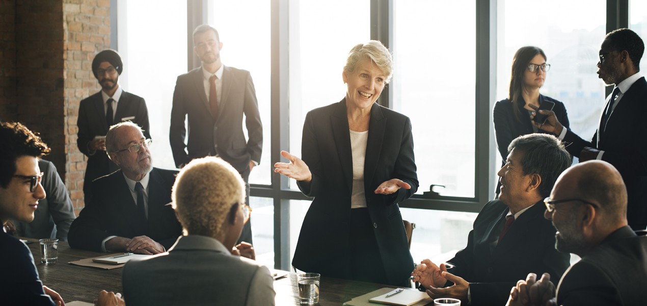 A team looking towards a mature woman standing and speaking during a meeting.