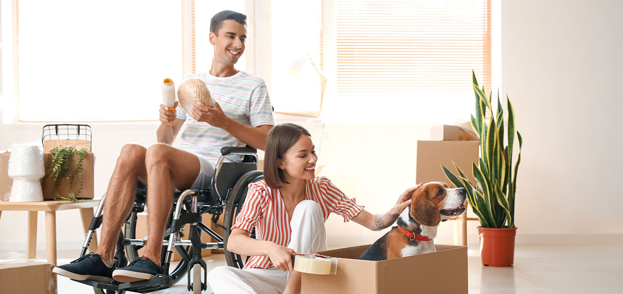 Young woman with her husband in wheelchair and dog packing things in a room on moving day.