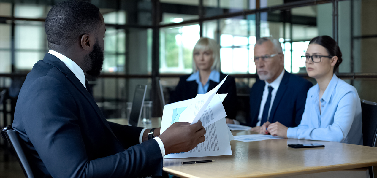 A business person holding papers in his hand while looking across at three business professionals who have serious expressions.