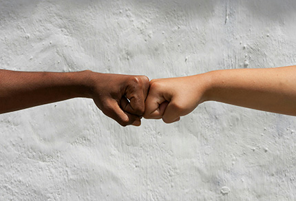 Two people of different races touching fists in front of a concrete wall.