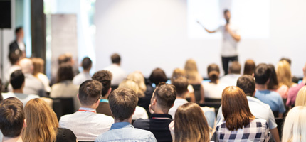 Back of an audience looking towards a speaker giving a talk at an event.
