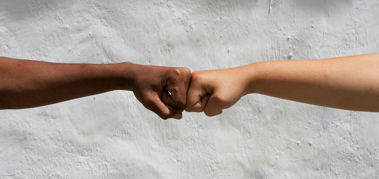 Two people of different races touching fists in front of a concrete wall.