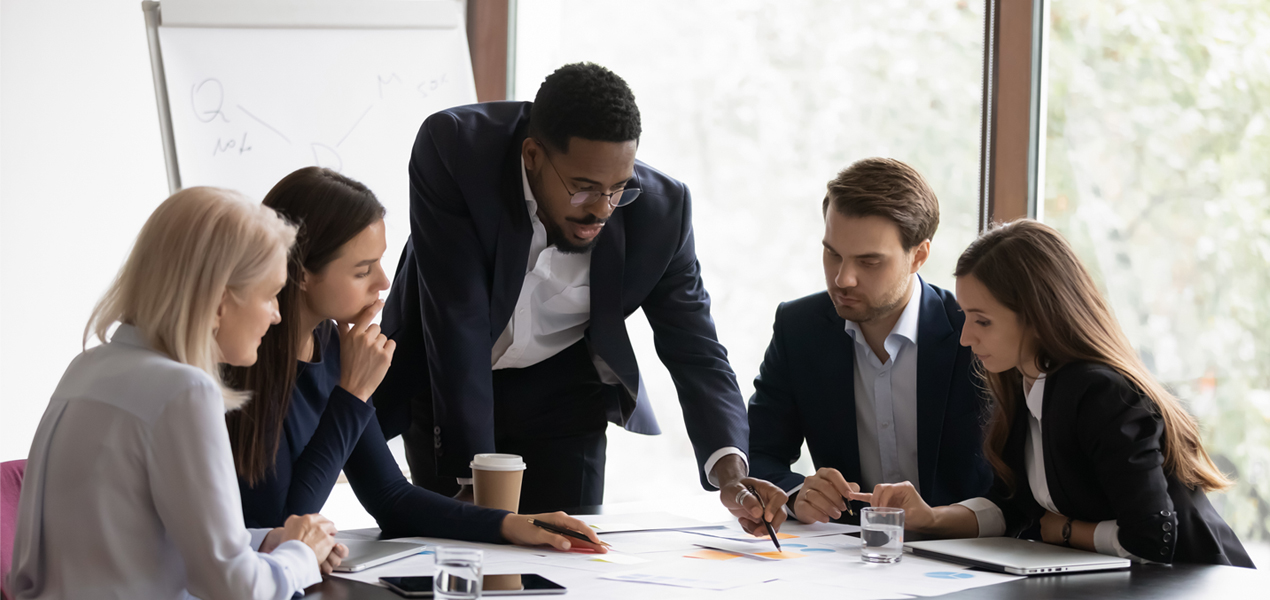 A diverse group of individuals having a meeting with one of them pointing to the papers in the middle of the table.