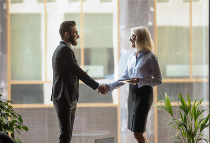 A business man and business woman shaking hands in front of a floor-to-ceiling window.