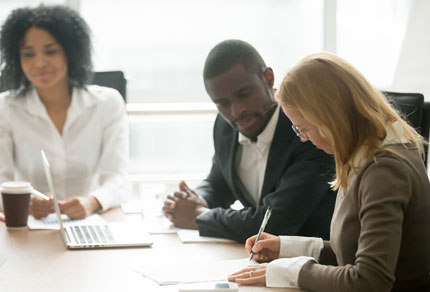 Three business people sat at a meeting table with one woman signing documents.