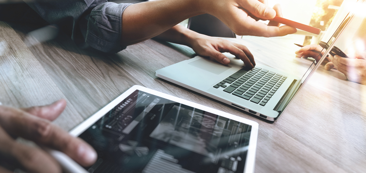 Close up of the hands of two people working side-by-side, one working on a tablet and one on a laptop.