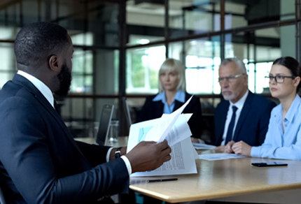 A business person holding papers in his hand while looking across at three business professionals who have serious expressions.
