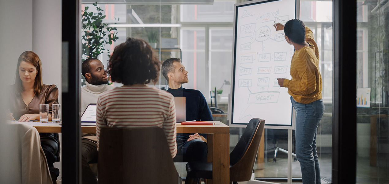 A group of business people sitting around a meeting table while a woman writes on a white board.