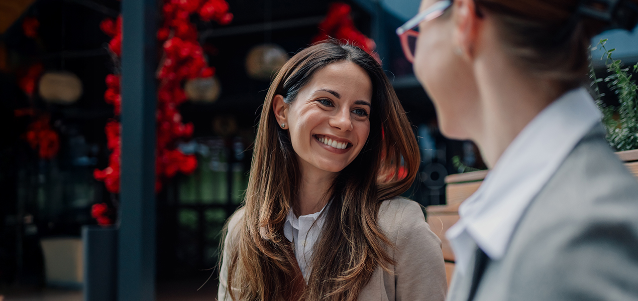 A smiling professional woman talking to another professional in an outdoor setting.