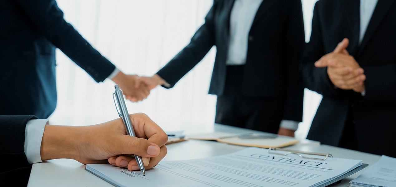 Close up on a person signing a contract with others shaking hands in the background.