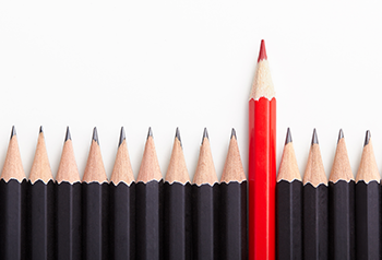A line of black pencil crayons with one red pencil crayon standing taller.